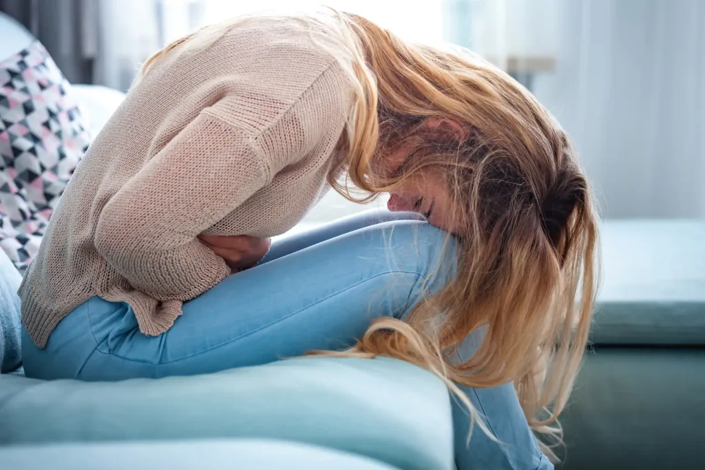 Young woman suffering from strong abdominal pain while sitting on sofa at home